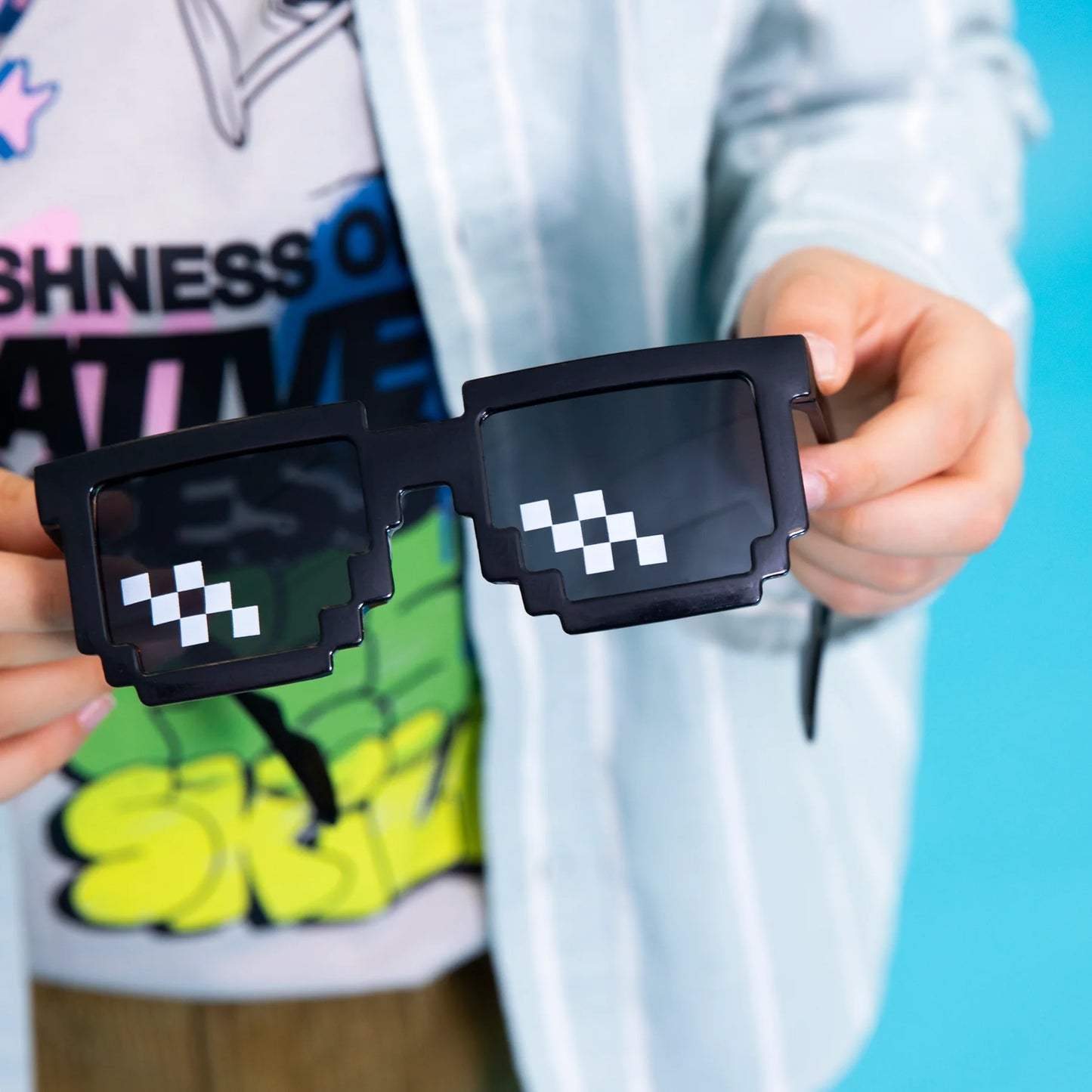 Ett par svarta pixel solglasögon med mörka linser och vita pixelrutor hålls i händerna mot färgglad bakgrund.